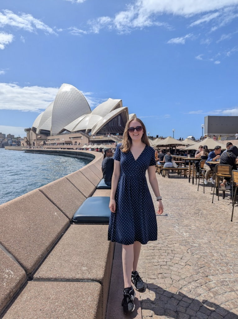 Woman in a navy dress standing in front of the Sydney Opera House on a sunny day.
